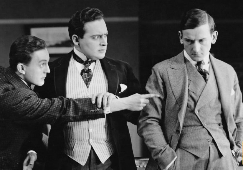 Three men in vintage attire engaged in a serious conversation indoors.